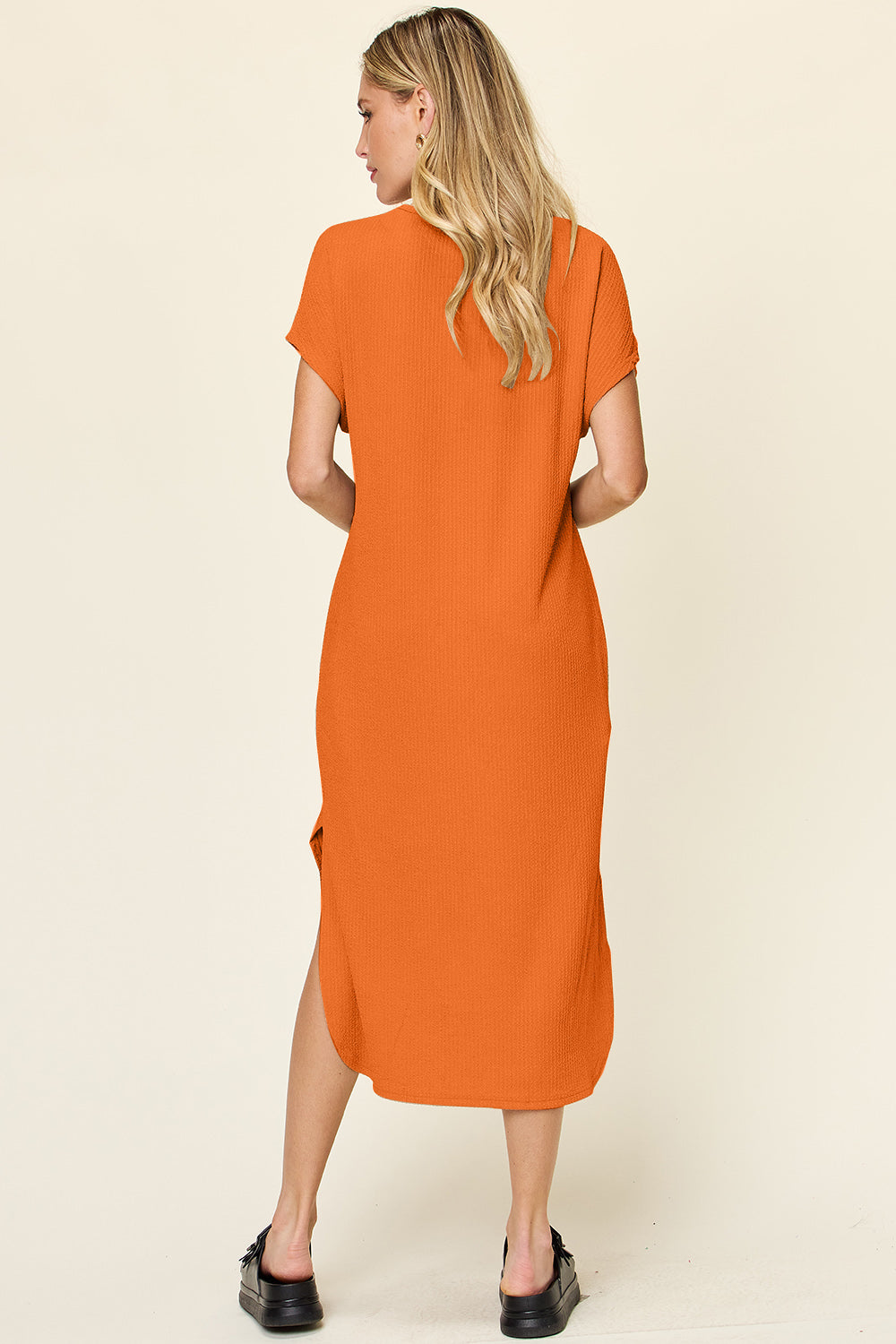 A person with long blonde hair wears a Double Take Full Size Round Neck Short Sleeve Slit Dress, an orange knee-length polyester spandex outfit, standing away from the camera. The dress features short sleeves, side slits, and is machine washable. They pair it with black shoes on a neutral background.