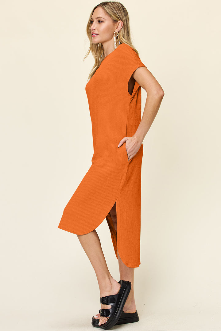 A woman poses in profile wearing a chic Double Take Full Size Round Neck Short Sleeve Slit Dress in orange. The knee-length, polyester spandex outfit is paired with black sandals. Her long blonde hair complements large hoop earrings against a plain background. This stylish dress is machine washable for easy care.
