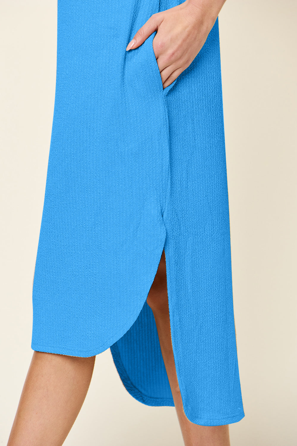 A person wears the Double Take Full Size Round Neck Short Sleeve Slit Dress, a blue, knee-length polyester spandex piece with textured fabric and a curved hem. The plus-size dress includes a pocket where the persons hand rests. The neutral background accentuates its elegant simplicity and machine washability.