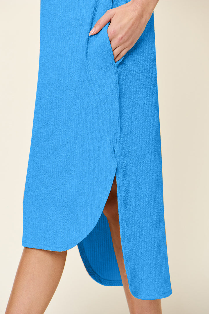 A person wears the Double Take Full Size Round Neck Short Sleeve Slit Dress, a blue, knee-length polyester spandex piece with textured fabric and a curved hem. The plus-size dress includes a pocket where the persons hand rests. The neutral background accentuates its elegant simplicity and machine washability.