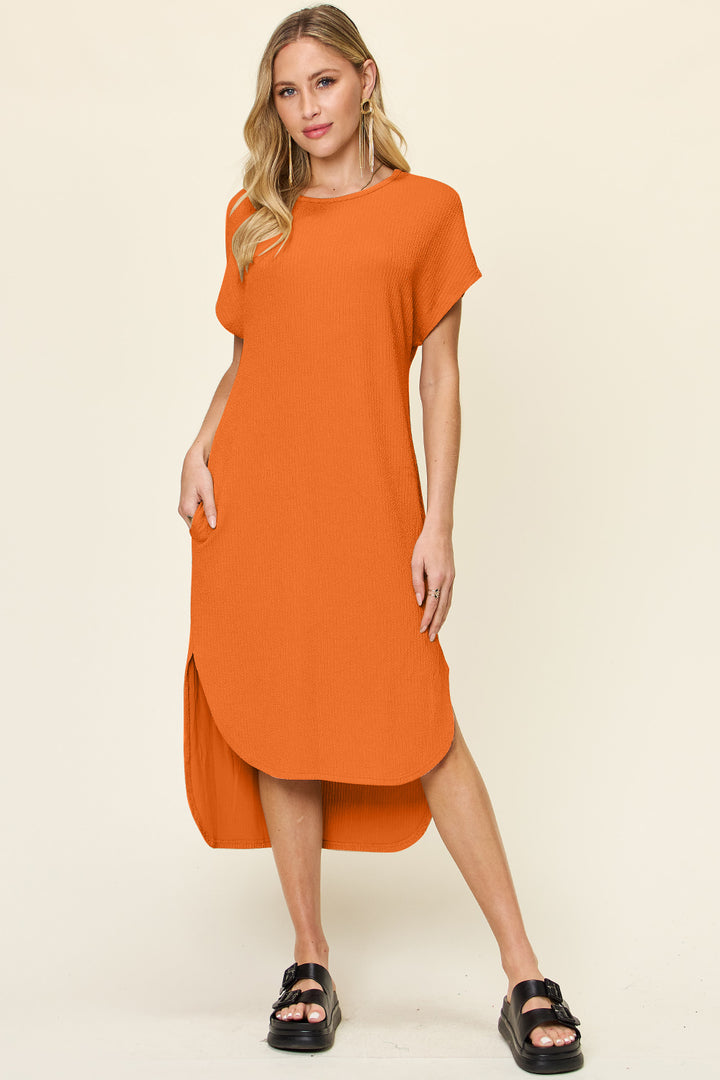A woman in a Double Take Full Size Round Neck Short Sleeve Slit Dress, machine washable and made from a polyester spandex blend, stands against a plain background. The loose orange dress with side slits complements her long blonde hair and black sandals. Her right hand is in her pocket, the other rests by her side.