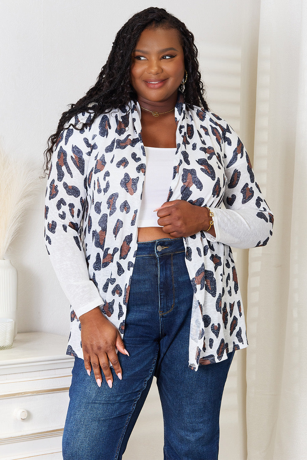 Standing in a well-lit room adorned with a white wall and decor, a person smiles, styled with long curls. They wear the trendy Double Take Leopard Long Sleeve Cardigan over a white top and blue jeans.