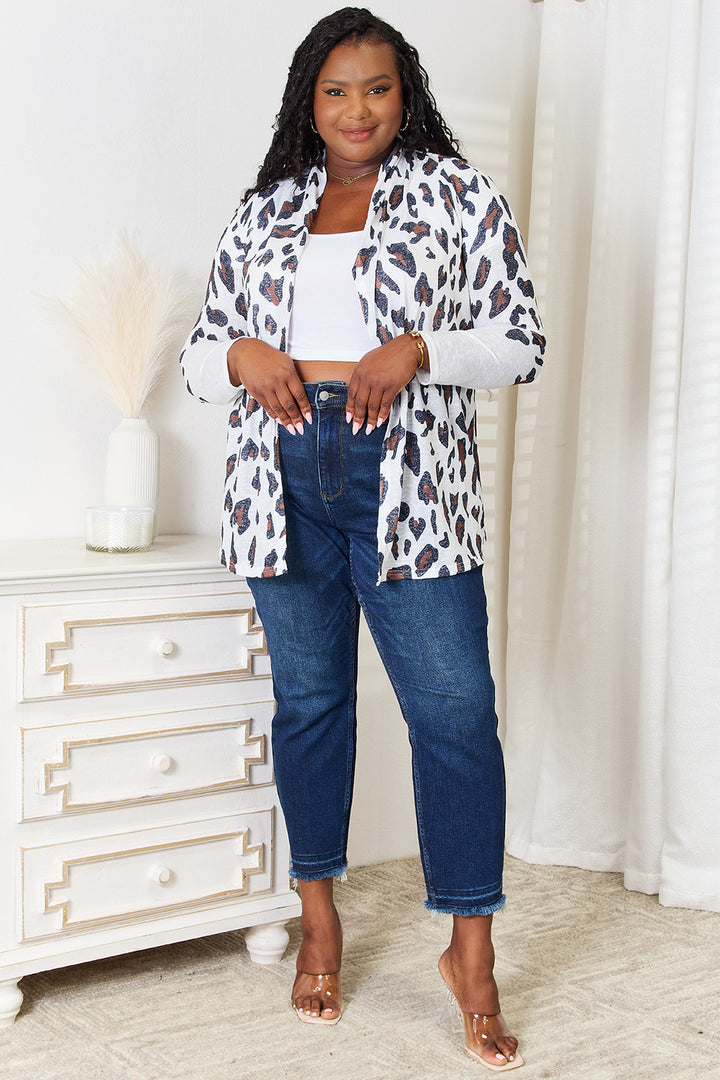 A woman in a stylish Double Take Leopard Long Sleeve Cardigan over a white top and dark blue jeans stands smiling in a room. She wears sandals, and beside her is a white chest of drawers adorned with decorative items.