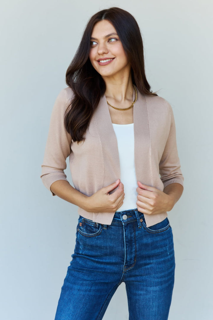 A person with long dark hair is smiling while glancing to the side, wearing the Doublju My Favorite Full Size 3/4 Sleeve Cropped Cardigan in Khaki over a white top, paired with blue jeans. This stylish cardigan perfectly complements the plain, light-colored background.