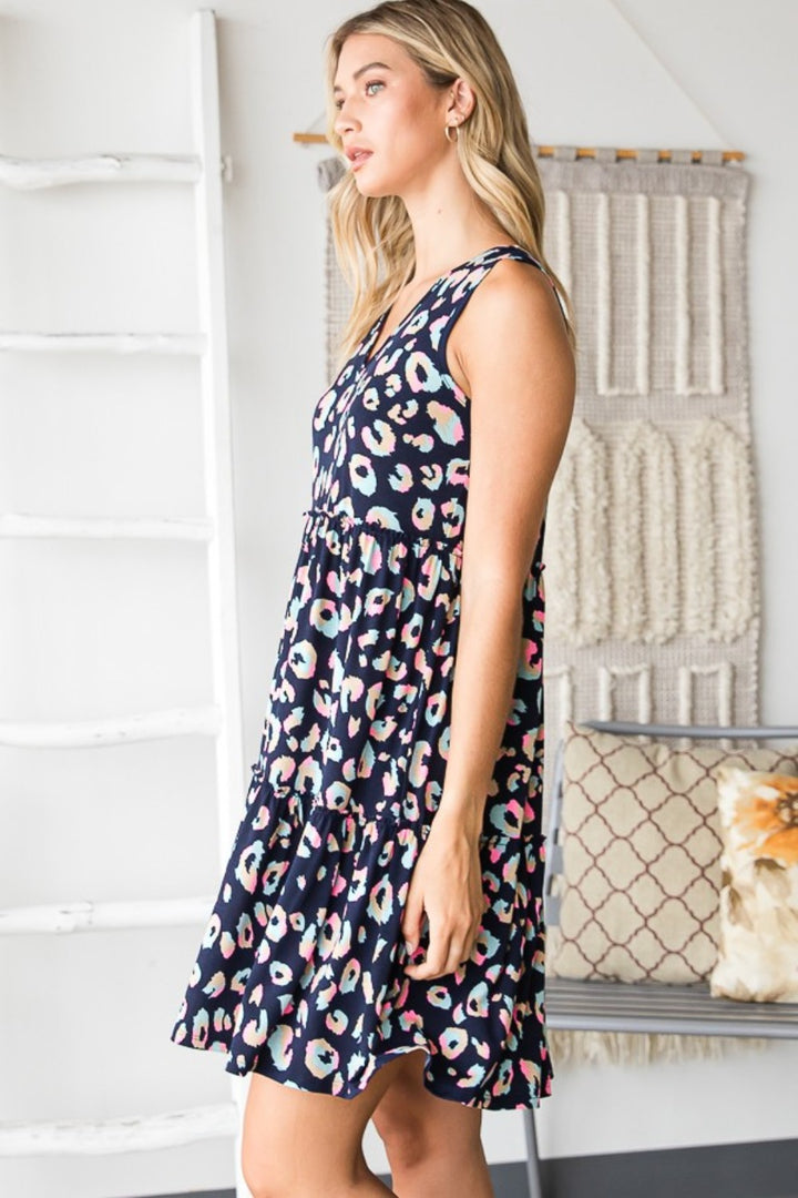 A woman wearing the Heimish Full Size Leopard V-Neck Ruffled Tank Dress is in a room featuring a decorative woven wall hanging and a cushioned bench. She gazes to the side, her long, light-colored hair flowing.