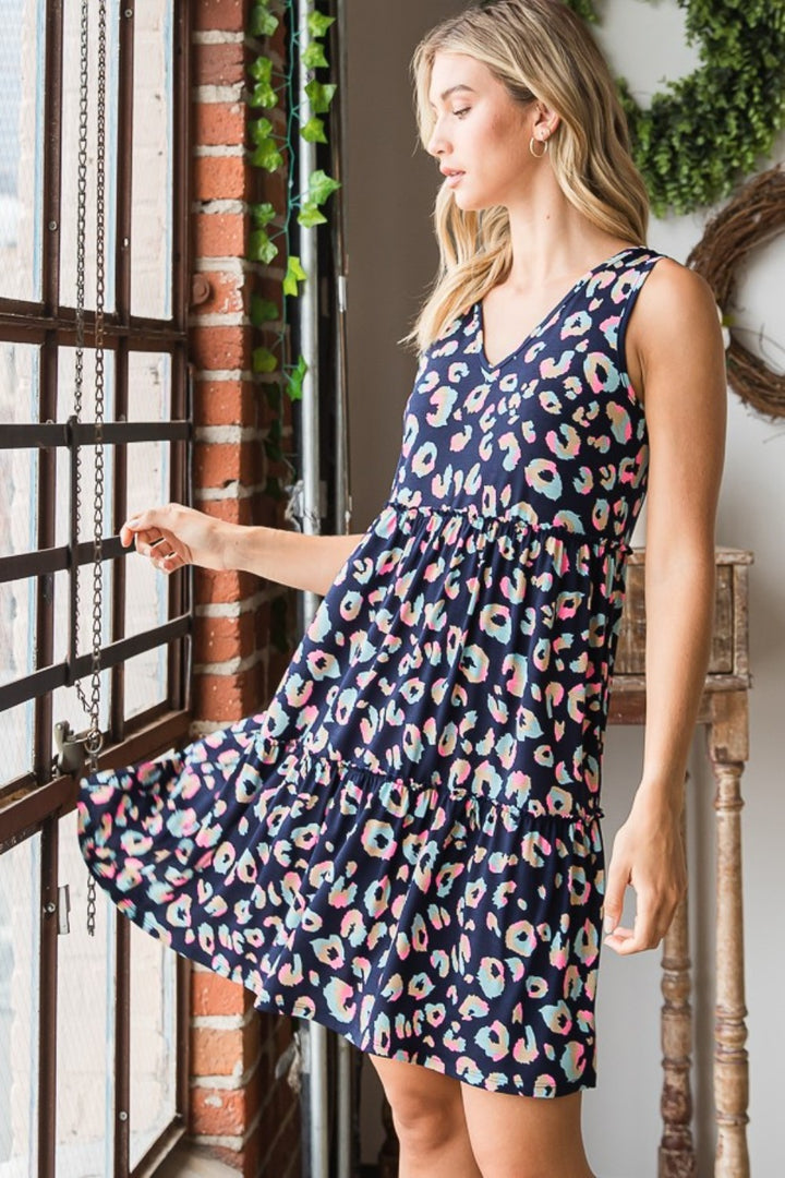 A woman in a Heimish Full Size Leopard V-Neck Ruffled Tank Dress stands by a large window, holding the hem and gazing out. The rooms brick walls and greenery provide an elegant contrast to her vibrant leopard print attire.