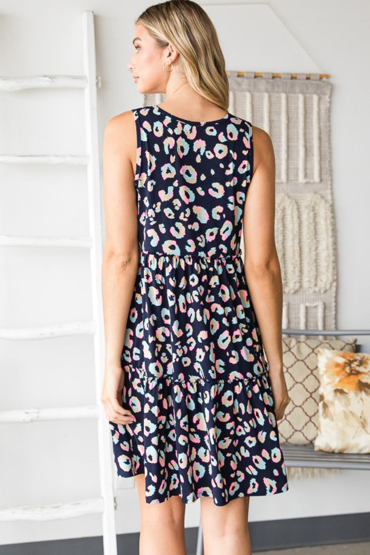 A woman in a Heimish Full Size Leopard V-Neck Ruffled Tank Dress stands gracefully in a room with white ladders and woven wall hangings. Her back faces the camera, adding mystery to her vibrant ensemble.