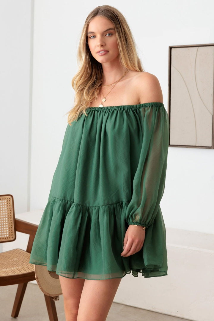 A woman with long blonde hair wears the stylish and chic Le Lis Organza Off Shoulder Puff Sleeve Mini Dress. She stands indoors next to a wooden chair, with minimalist beige wall art as the backdrop.