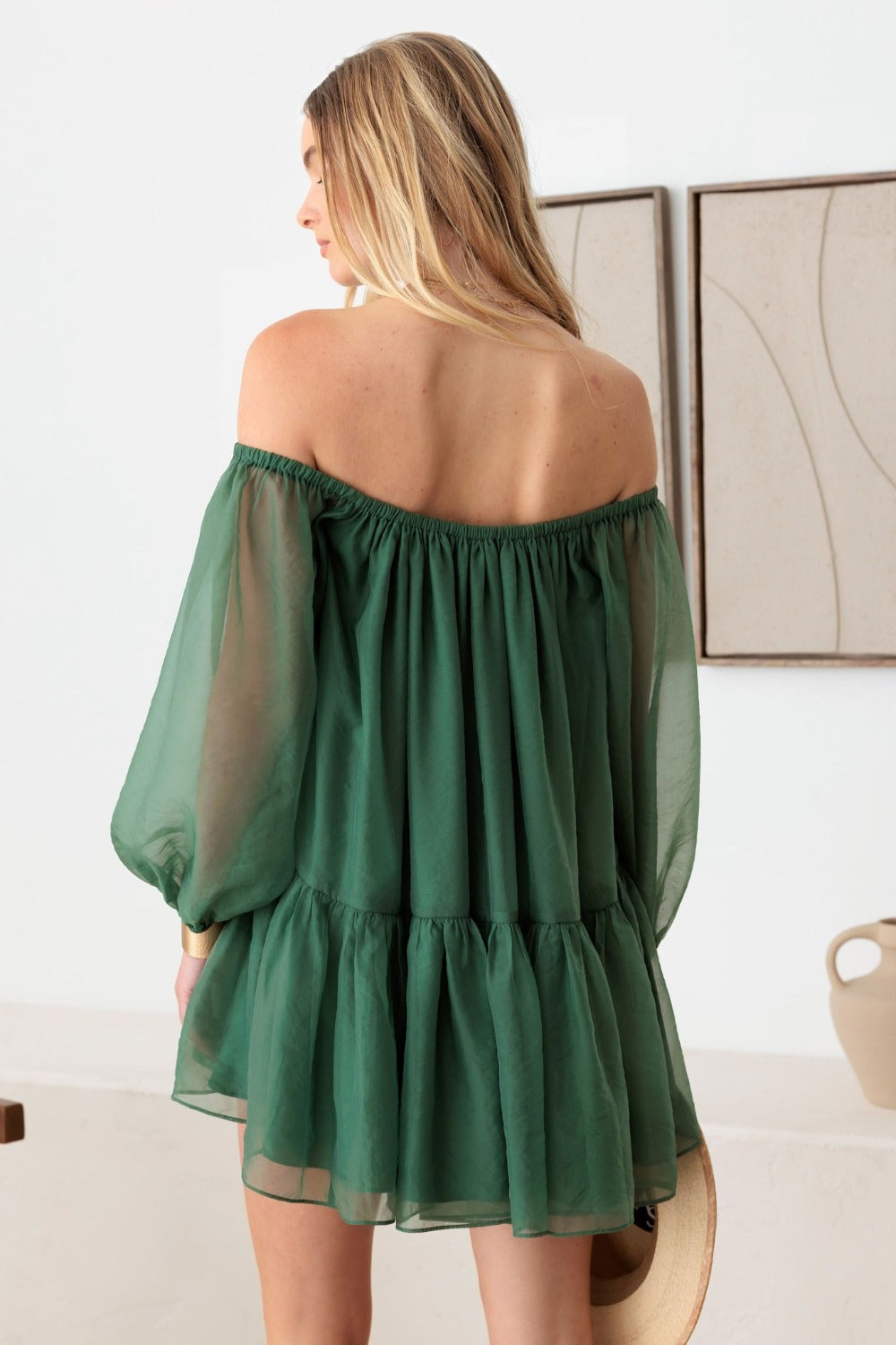 A woman with long blonde hair is seen from the back, dressed in the stylish and chic Le Lis Organza Off Shoulder Puff Sleeve Mini Dress. She holds a straw hat by her side, set against a backdrop of abstract wall art and a minimalist interior.