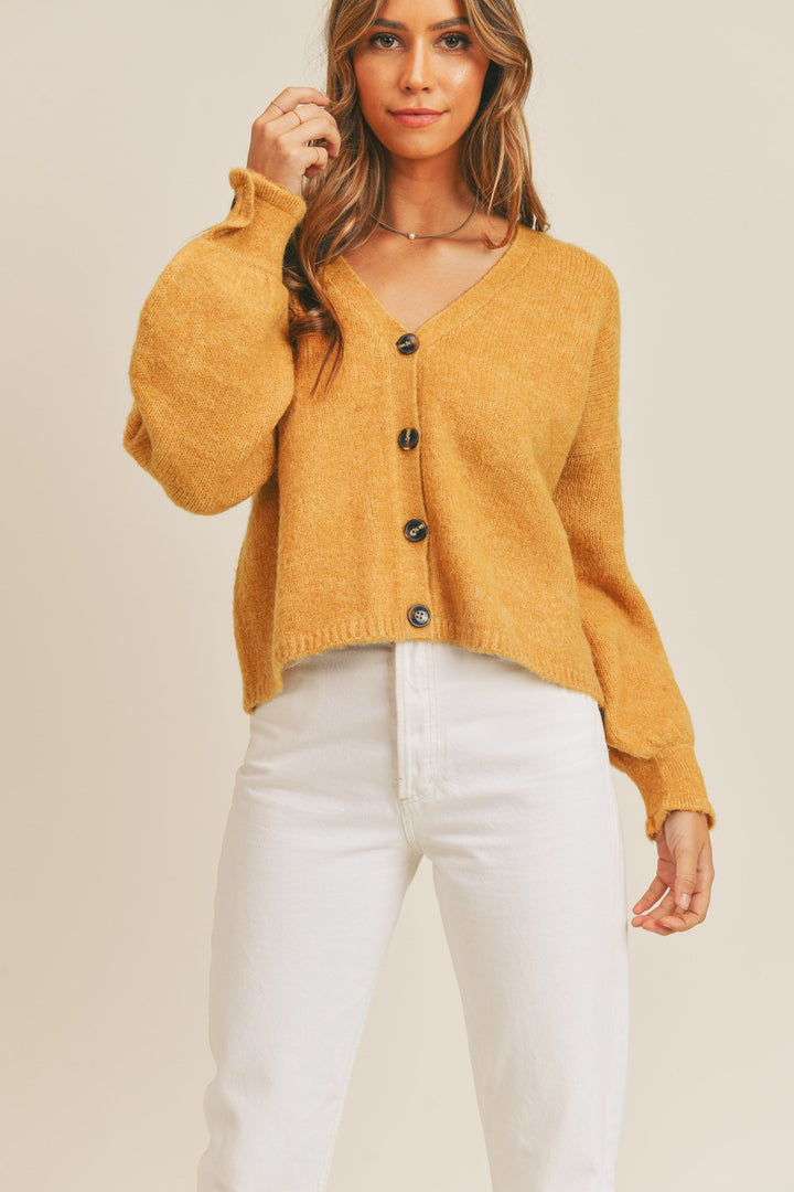 A person wearing a mustard yellow MABLE Long Sleeve Button Down Sweater Cardigan and white pants poses against a neutral background. The knit fabric cardigan, featuring a V-neck and balloon sleeves, adds a touch of cozy elegance.