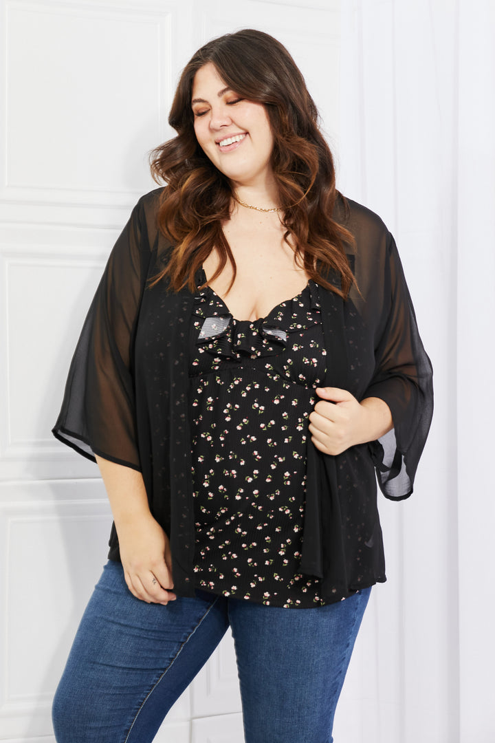 A woman with long brown hair smiles while wearing the Melody Just Breathe Full Size Chiffon Kimono in black over a floral top and blue jeans. She exudes casual chic as she stands against a light-colored wall, holding the kimono with one hand.