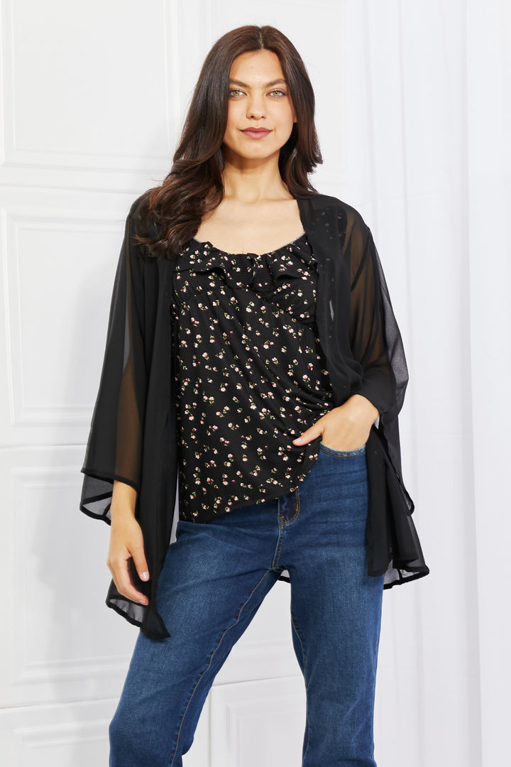 A person with long dark hair in the Melody Just Breathe Full Size Chiffon Kimono in Black stands against a white background, exuding casual chic. The kimono's floral pattern and sheer fabric accentuate their look as they pair it with blue jeans, showcasing one hand in their pocket for a relaxed yet elegant touch.