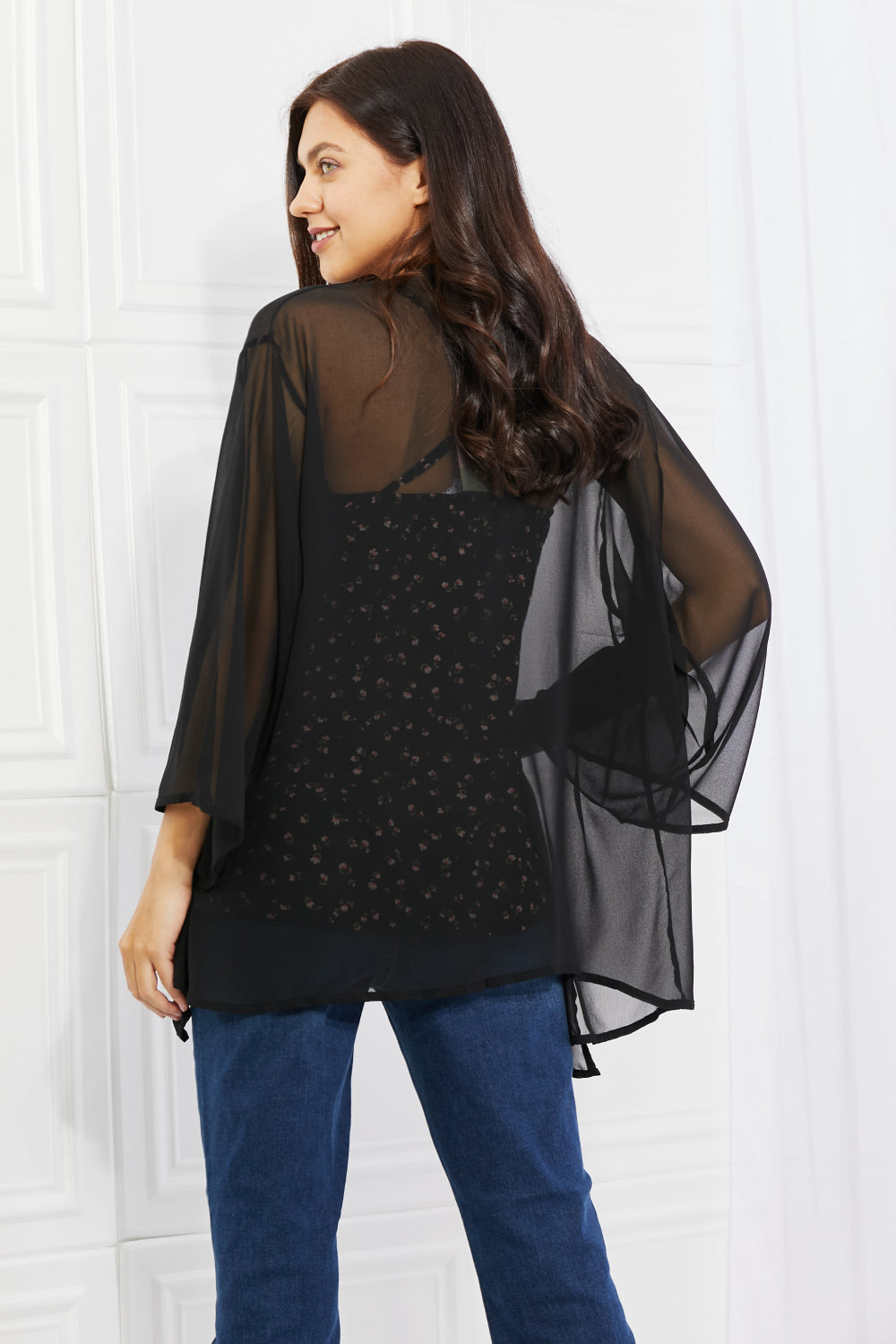 A woman with long dark hair stands facing away, showcasing an effortlessly chic look in the Melody Just Breathe Full Size Chiffon Kimono in black, featuring flowing sleeves. She pairs it with a floral-patterned shirt and blue jeans, all set against the backdrop of a white room with paneled walls.