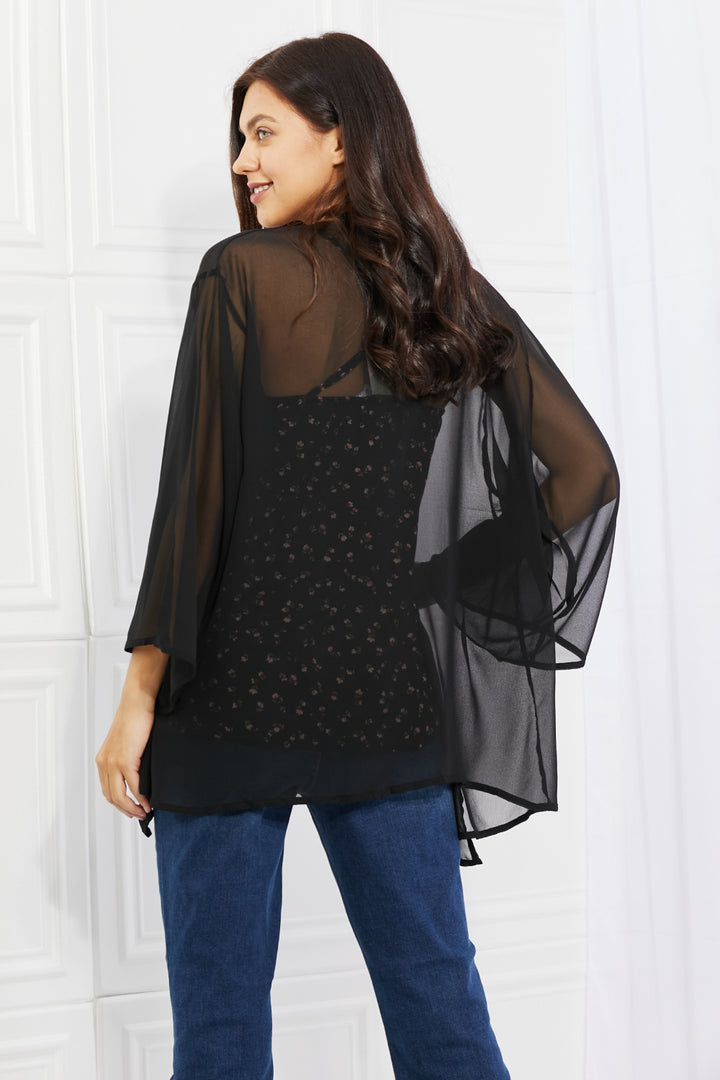 A woman with long dark hair stands facing away, showcasing an effortlessly chic look in the Melody Just Breathe Full Size Chiffon Kimono in black, featuring flowing sleeves. She pairs it with a floral-patterned shirt and blue jeans, all set against the backdrop of a white room with paneled walls.