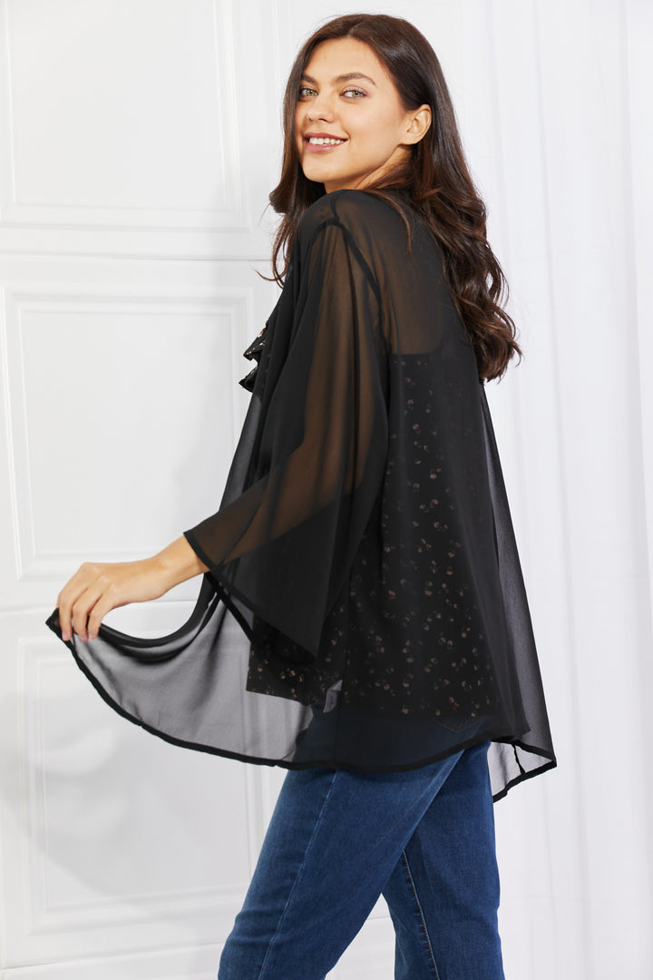A woman exudes casual chic as she smiles in the Melody Just Breathe Full Size Chiffon Kimono in Black, complete with elegant kimono-style sleeves. Her long dark hair complements the patterned black top and blue jeans, all set against a white paneled background.