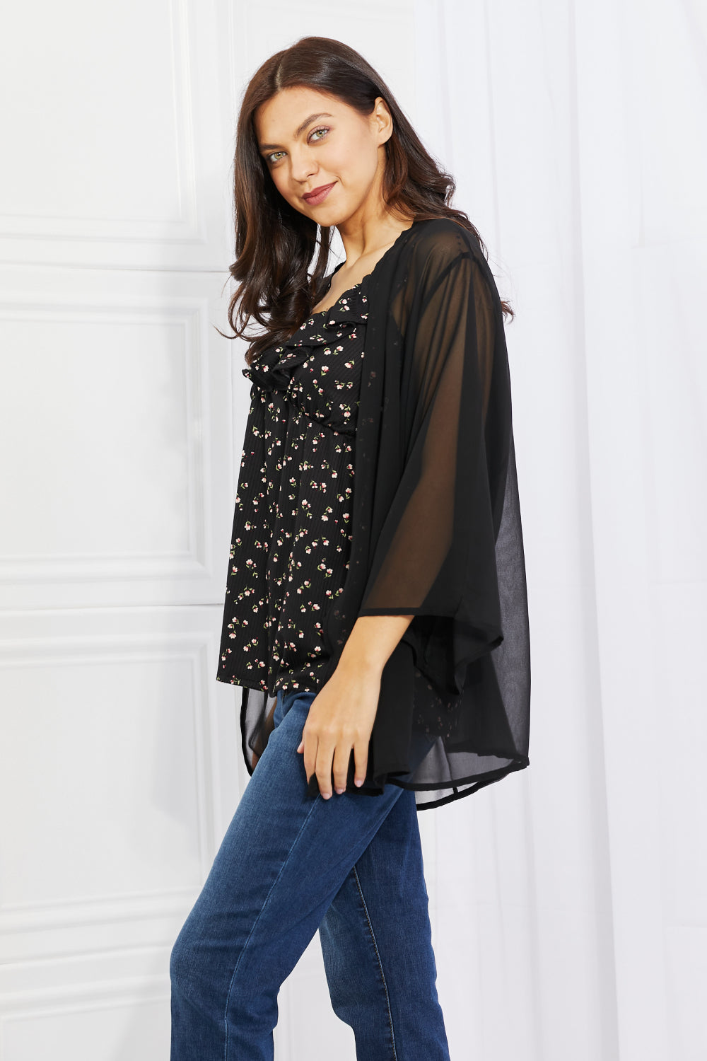 A person with long hair is wearing a black floral top and the Melody Just Breathe Full Size Chiffon Kimono in Black, paired with blue jeans. They are standing in a bright room with a white wall, smiling slightly, and looking towards the camera, exuding casual chic vibes.