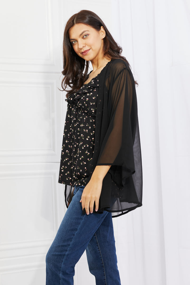 A person with long hair is wearing a black floral top and the Melody Just Breathe Full Size Chiffon Kimono in Black, paired with blue jeans. They are standing in a bright room with a white wall, smiling slightly, and looking towards the camera, exuding casual chic vibes.