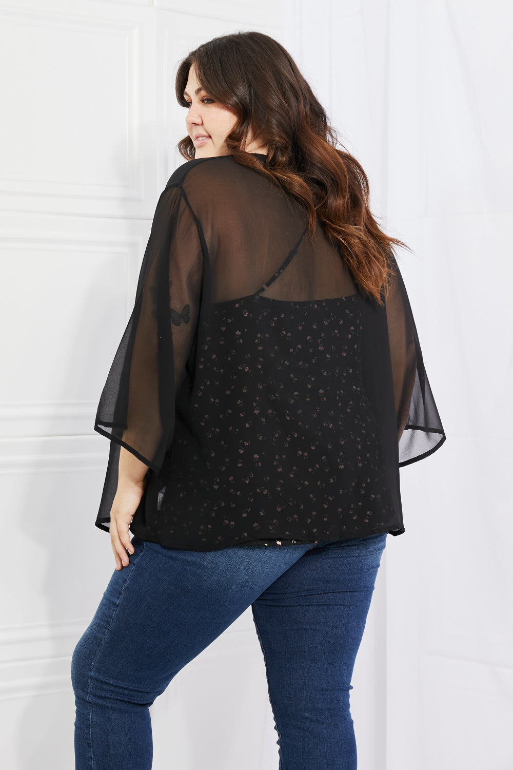 A person with long hair is standing and facing away, wearing the Melody Just Breathe Full Size Chiffon Kimono in Black over a patterned top and blue jeans. The setting appears to be indoors with a light-colored wall, exuding a casual chic vibe.
