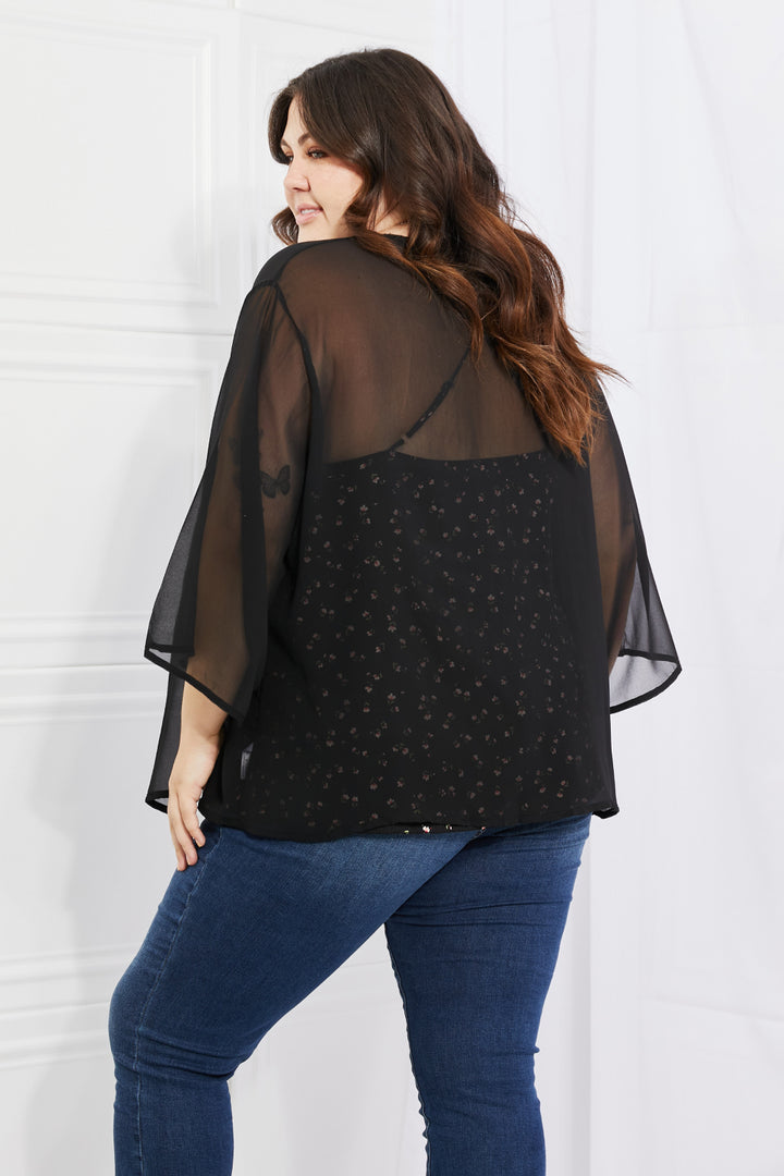 A person with long hair is standing and facing away, wearing the Melody Just Breathe Full Size Chiffon Kimono in Black over a patterned top and blue jeans. The setting appears to be indoors with a light-colored wall, exuding a casual chic vibe.