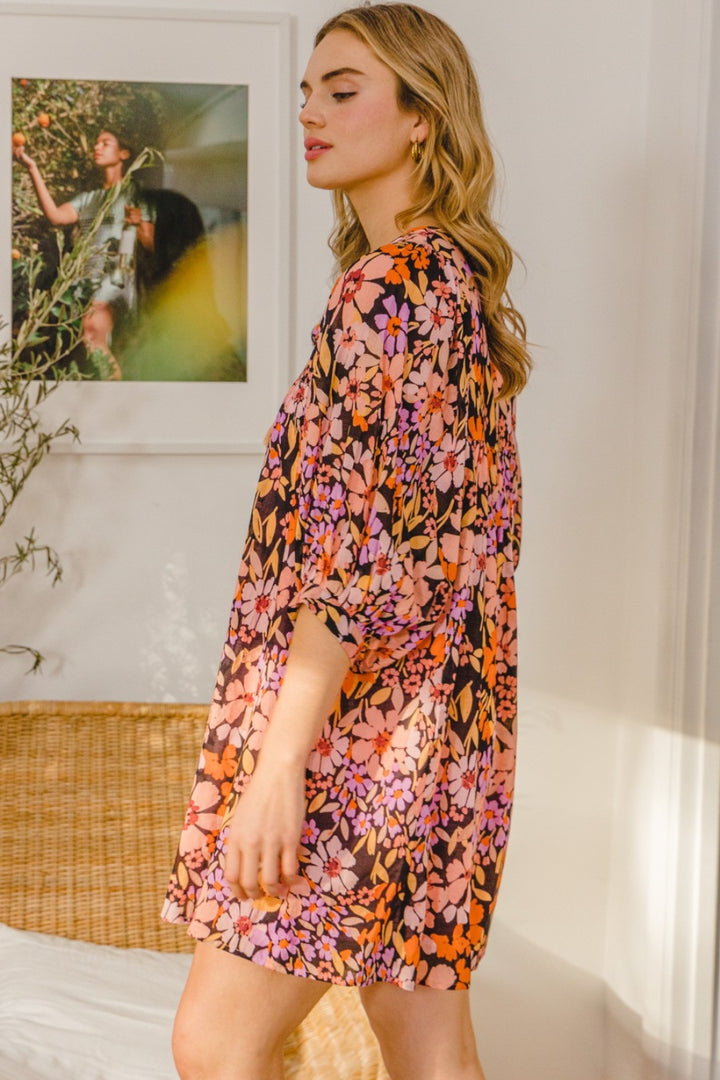 A woman with wavy blonde hair wears the ODDI Full Size Floral Tied Neck Mini Dress in pink, orange, and black floral print, standing indoors near a rattan bench and framed artwork on the wall.