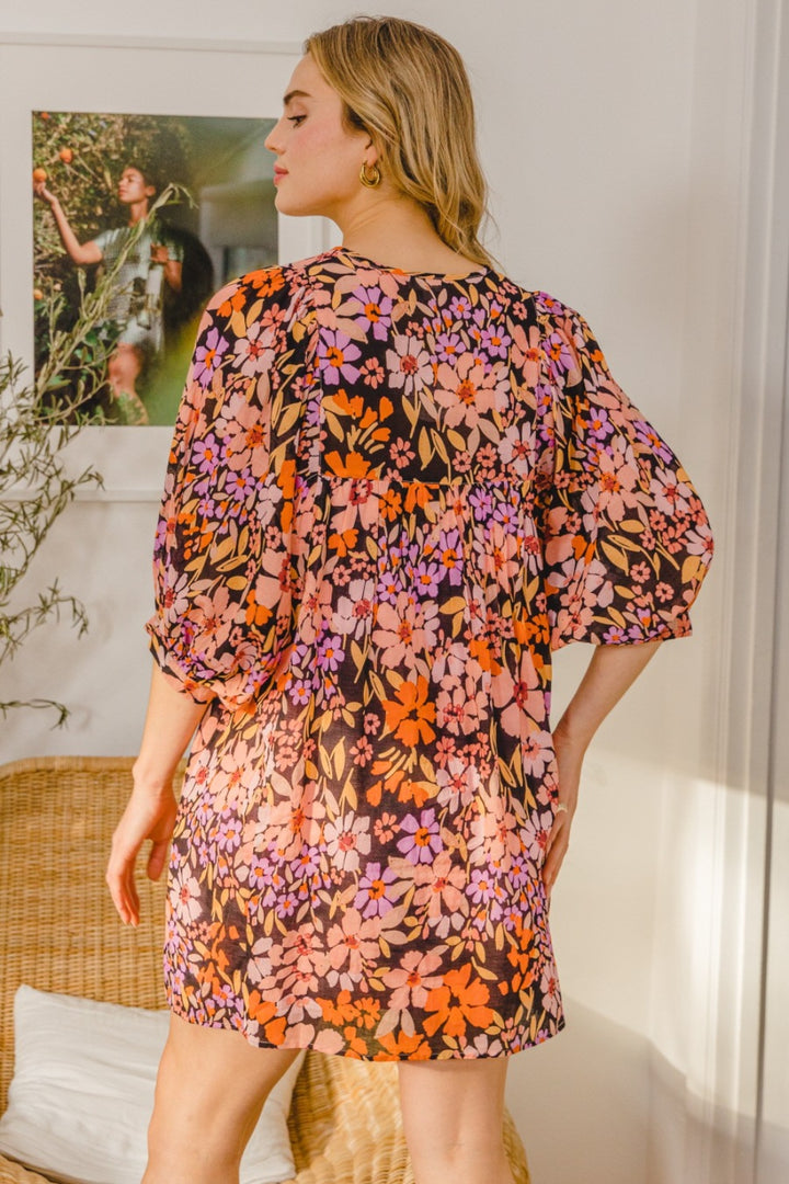 A woman stands indoors with her back to the camera, wearing an ODDI Full Size Floral Tied Neck Mini Dress in bright orange, pink, and purple. The scene features wicker furniture and a framed photo on the wall.