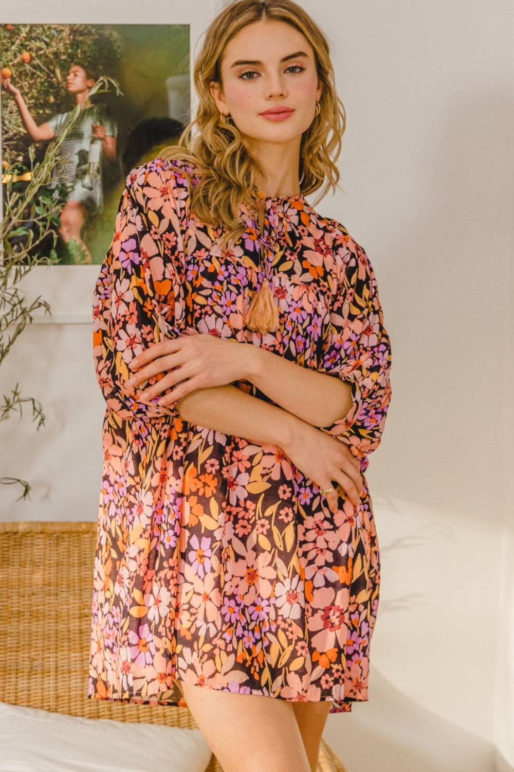 A woman stands indoors, wearing the ODDI Full Size Floral Tied Neck Mini Dress featuring a vibrant pink, orange, purple, and black floral pattern. She poses with crossed arms; a wicker chair and plant are in the background.