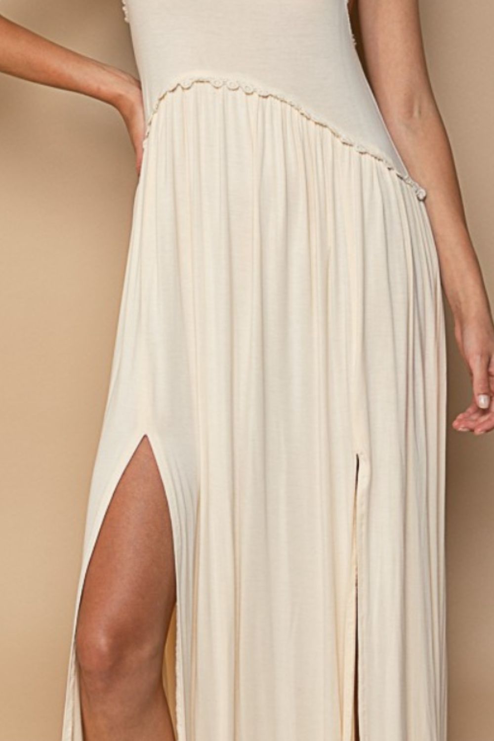 Against a tan background, a woman exudes confidence in the POL Sleeveless Back Zipper Front Slit Maxi Dress. The cream knit maxi dress, featuring a racerback and curved waistline with subtle trim, is styled with one hand on her hip, highlighting her poised elegance.