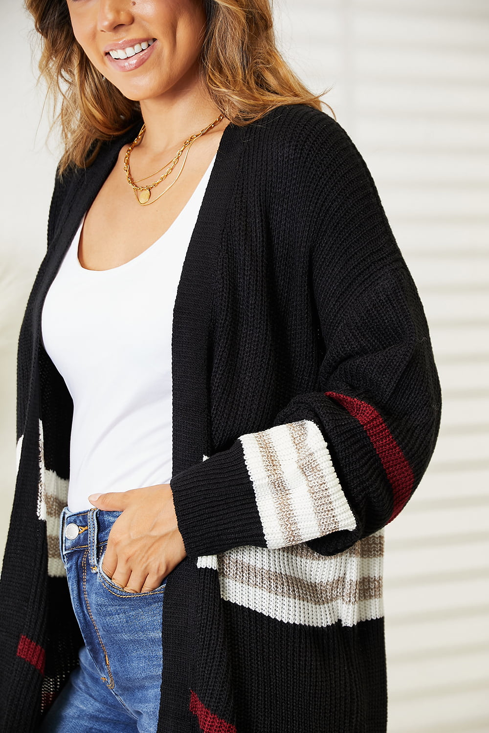 A person wearing the Perfee Striped Rib-Knit Drop Shoulder Open Front Cardigan, featuring red, white, and beige accents on the sleeves, over a white top and blue jeans. They complement their look with a layered gold necklace and are smiling against a light-colored background.