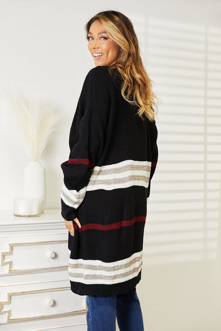 A woman with long hair smiles while wearing the Perfee Striped Rib-Knit Drop Shoulder Open Front Cardigan, featuring white and red accents. She stands near a white dresser adorned with dried plants, leaning slightly back as sunlight casts shadows on the wall beside her.