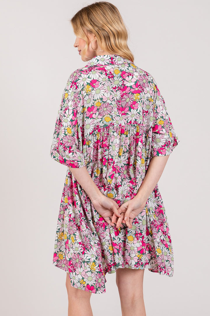 A person with blonde hair stands with their back to the camera, wearing a SAGE + FIG Floral Button Down Mini Shirt Dress featuring pink, white, and yellow flowers with green leaves. Their hands are clasped behind their back, adding an air of mystery to this charming scene.