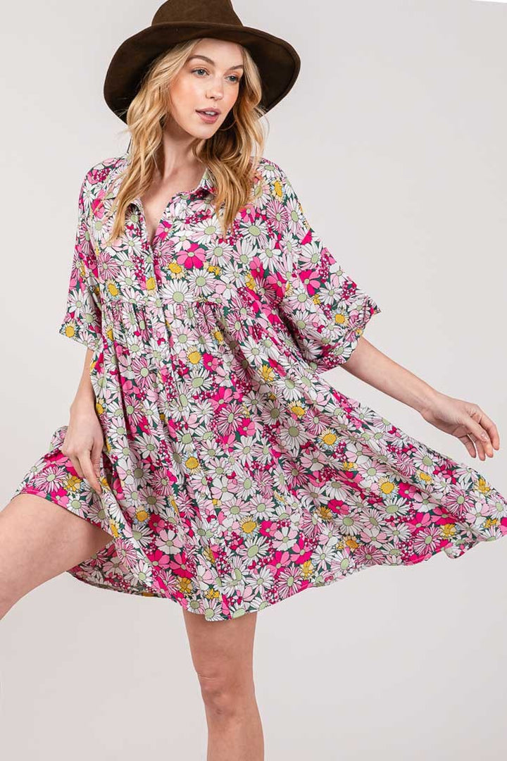 Wearing a SAGE + FIG Floral Button Down Mini Shirt Dress, a person gracefully poses with a wide-brimmed brown hat. They stand with one leg slightly raised, holding out the dress to create a flowing effect against the plain gray background.