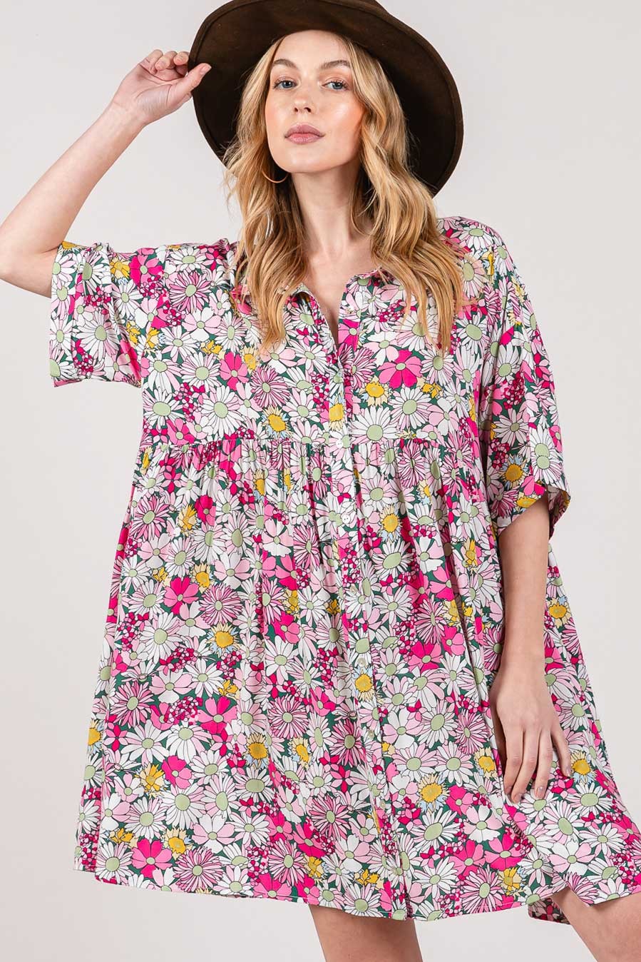 A woman wearing a wide-brimmed hat and the SAGE + FIG Floral Button Down Mini Shirt Dress, adorned with pink, white, and yellow flowers in a colorful pattern, poses with one hand on the hats brim against a plain background.