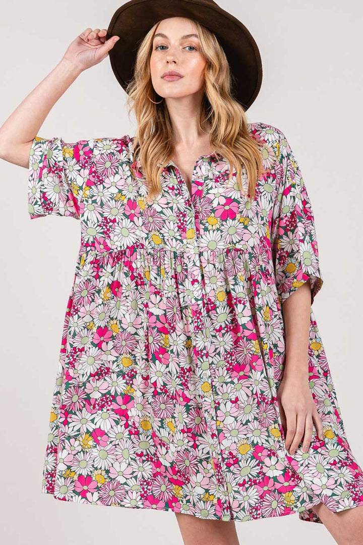 A woman wearing a wide-brimmed hat and the SAGE + FIG Floral Button Down Mini Shirt Dress, adorned with pink, white, and yellow flowers in a colorful pattern, poses with one hand on the hats brim against a plain background.