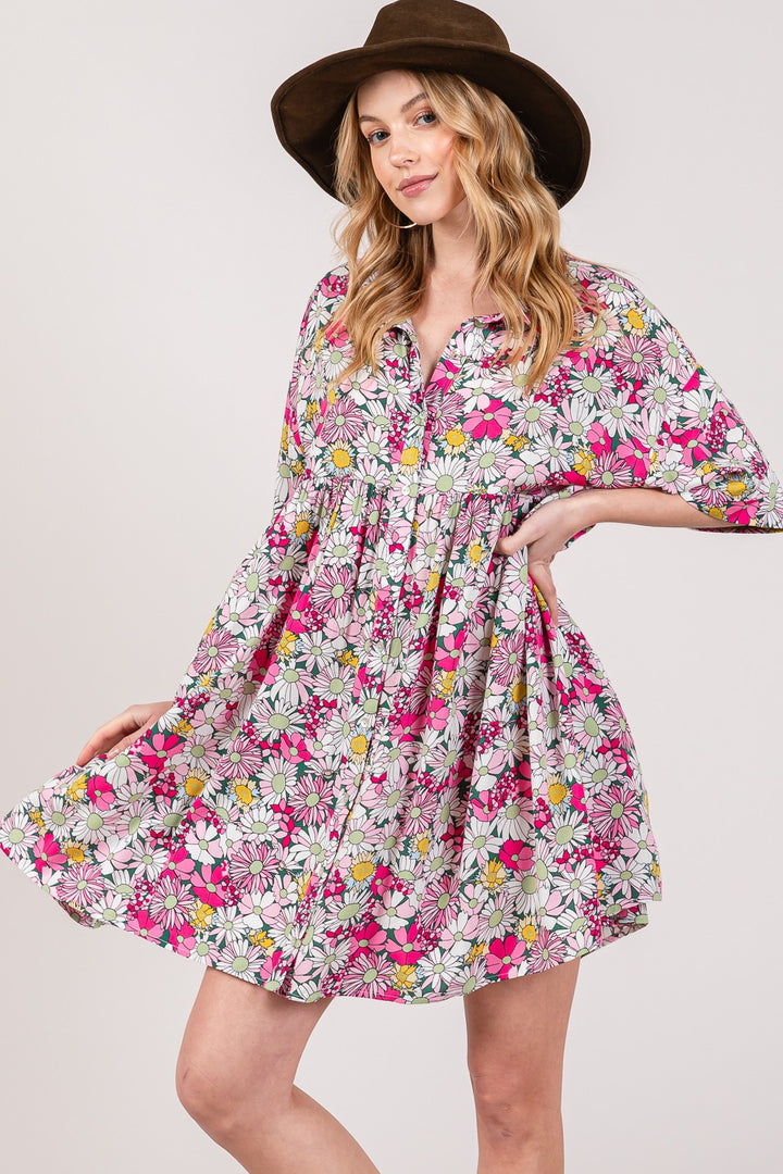 A person wearing a wide-brimmed brown hat and the SAGE + FIG Floral Button Down Mini Shirt Dress with vibrant pink, yellow, and white flowers. The loose fit beautifully complements their long, wavy hair as they confidently pose with one hand on their hip.
