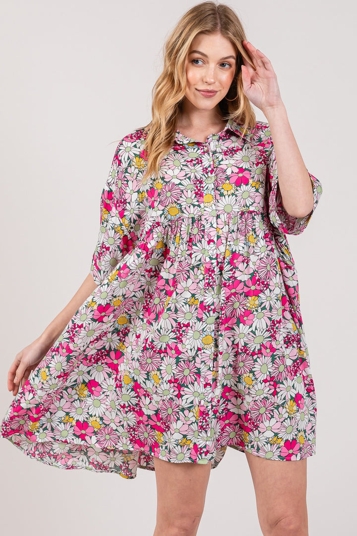 A woman in a SAGE + FIG Floral Button Down Mini Shirt Dress smiles against a plain background. Shes holding out one side of the pink dress, highlighting its flowy design adorned with white, pink, and yellow flowers.