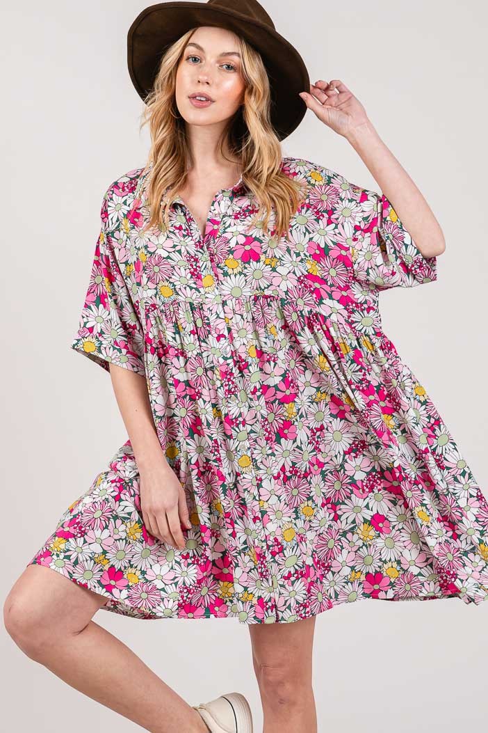 A woman in a wide-brimmed hat poses with one hand on its brim and one knee slightly bent, wearing the SAGE + FIG Floral Button Down Mini Shirt Dress adorned with pink, yellow, and white flowers on a light background against a plain backdrop.