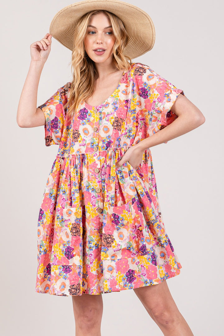 Someone poses in a wide-brimmed straw hat and the SAGE + FIG Floral Short Sleeve Babydoll Dress with Pockets, placing one hand on the hat and the other on their hip. The dress is loose-fitting, knee-length, features short sleeves, and has hidden pockets for extra charm and functionality.
