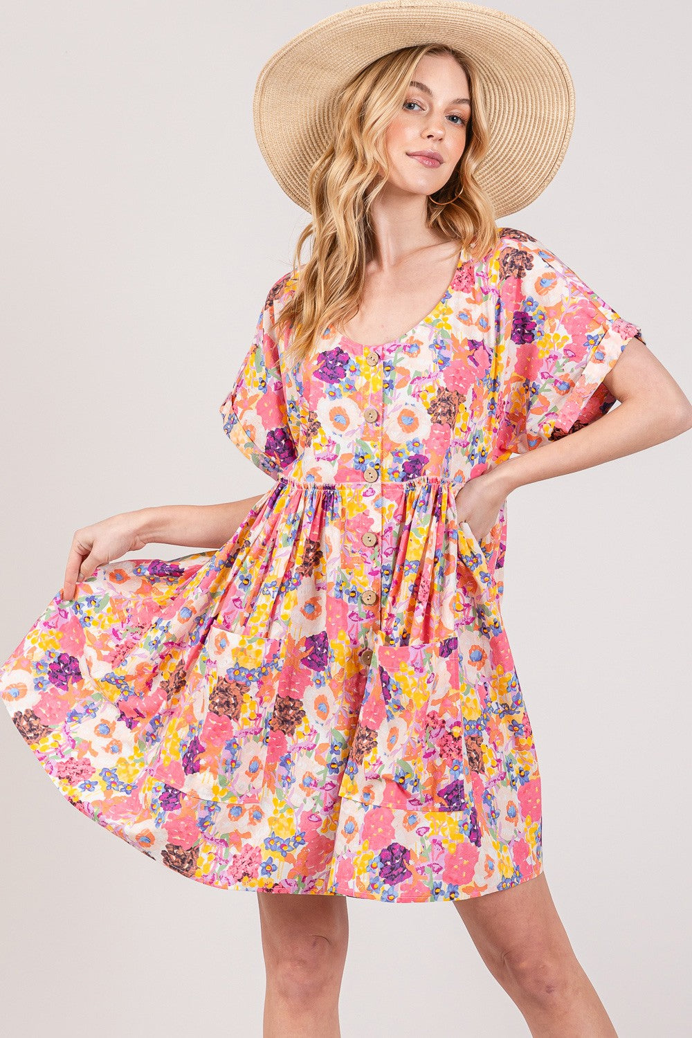 A woman poses confidently against a plain background in a SAGE + FIG Floral Babydoll Dress with Pockets, featuring vibrant pink, yellow, and purple hues. Wearing a wide-brimmed straw hat, she gently smiles while holding her dress out to the side.