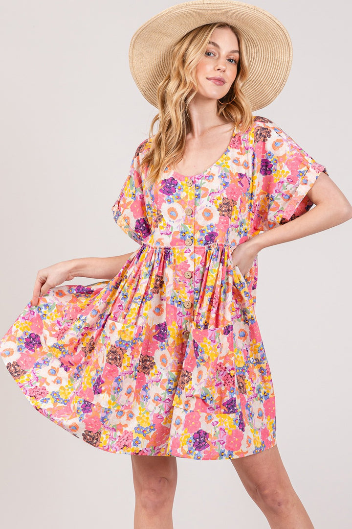 A woman poses confidently against a plain background in a SAGE + FIG Floral Babydoll Dress with Pockets, featuring vibrant pink, yellow, and purple hues. Wearing a wide-brimmed straw hat, she gently smiles while holding her dress out to the side.