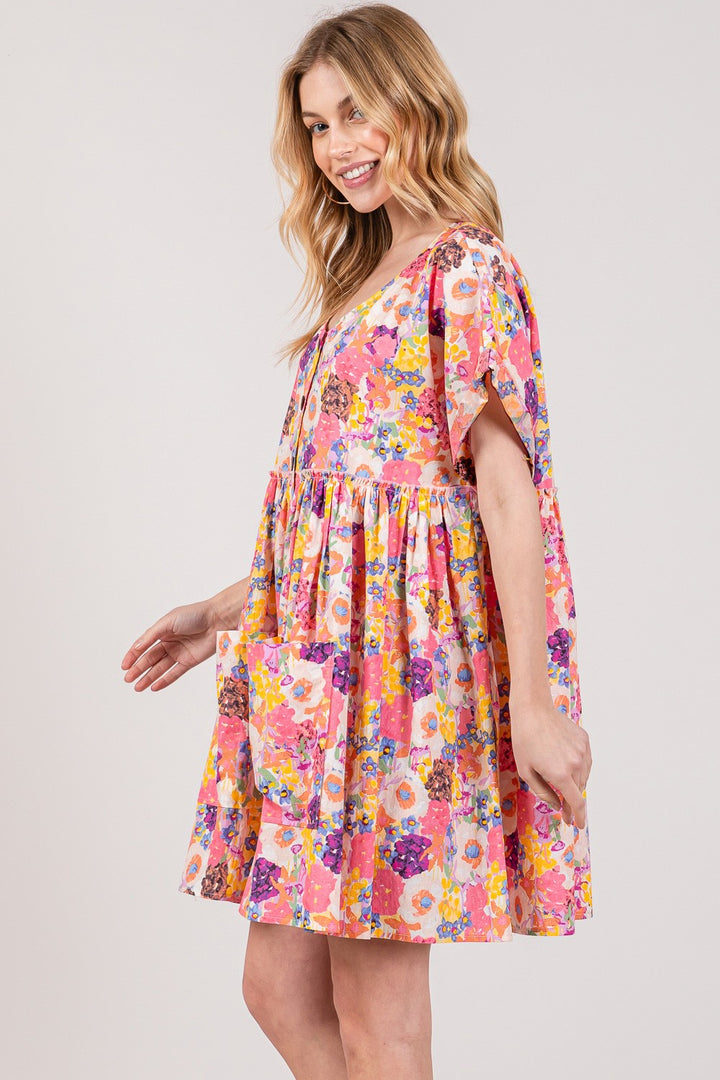 A smiling person with long hair wears the SAGE + FIG Floral Short Sleeve Babydoll Dress, featuring a loose skirt and large pockets. They stand slightly turned against a plain background.