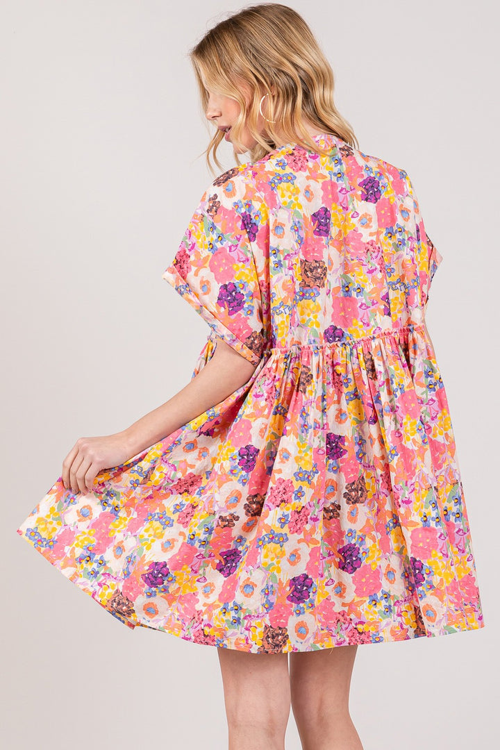 A blonde woman stands sideways in a SAGE + FIG Floral Short Sleeve Babydoll Dress with Pockets, holding the hem to display its flowy design. The plain, neutral background enhances the charm of her colorful, loose-fitting dress.