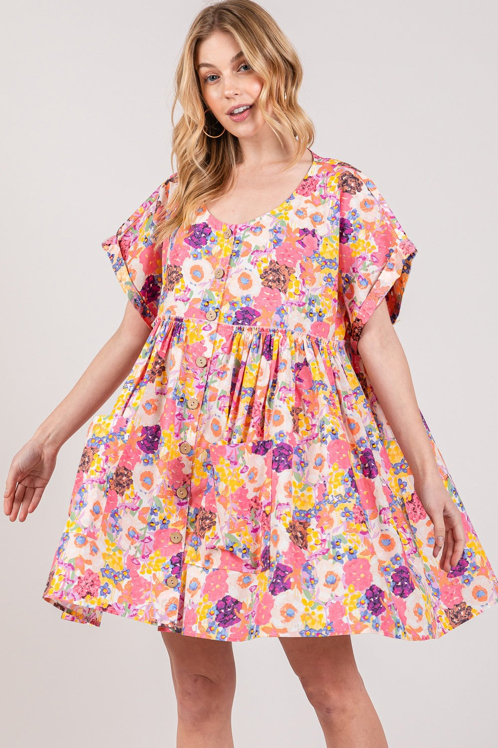 A smiling woman is wearing the SAGE + FIG Floral Short Sleeve Babydoll Dress with Pockets, showcasing a loose fit with vibrant pink, yellow, and purple floral patterns on a plain white background.