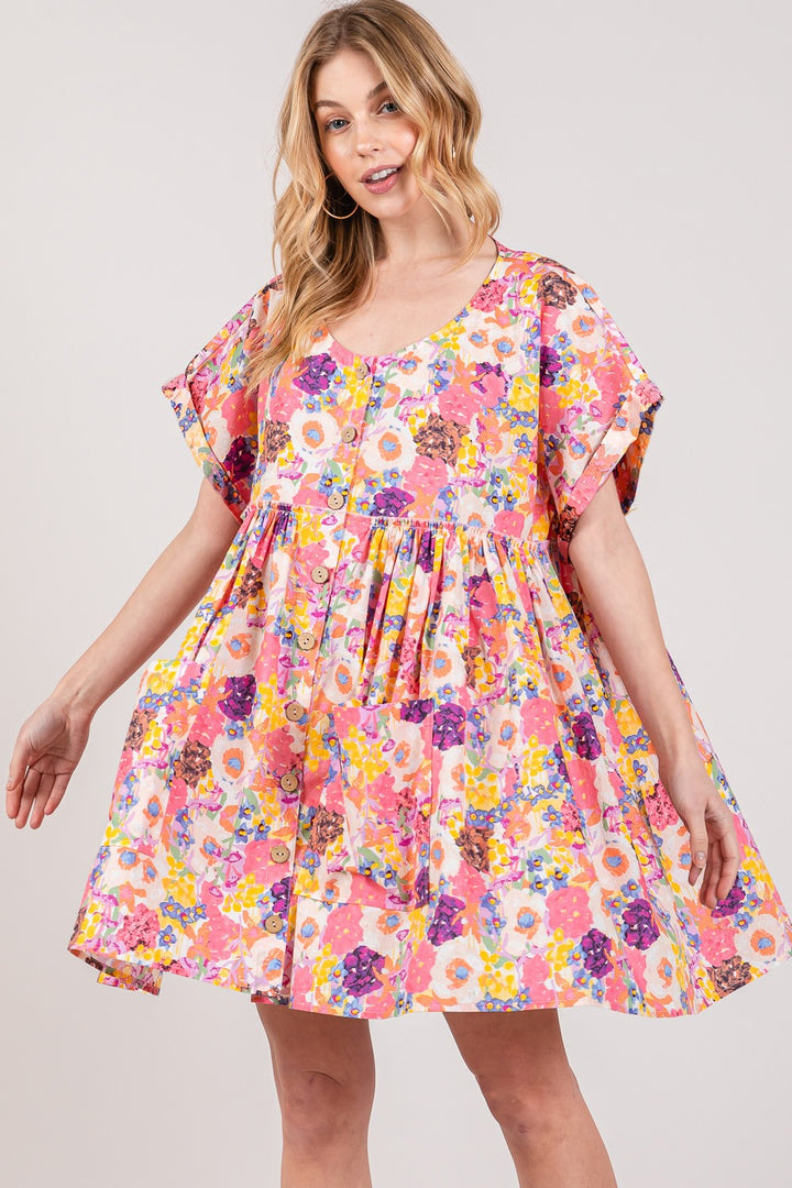 A smiling woman is wearing the SAGE + FIG Floral Short Sleeve Babydoll Dress with Pockets, showcasing a loose fit with vibrant pink, yellow, and purple floral patterns on a plain white background.