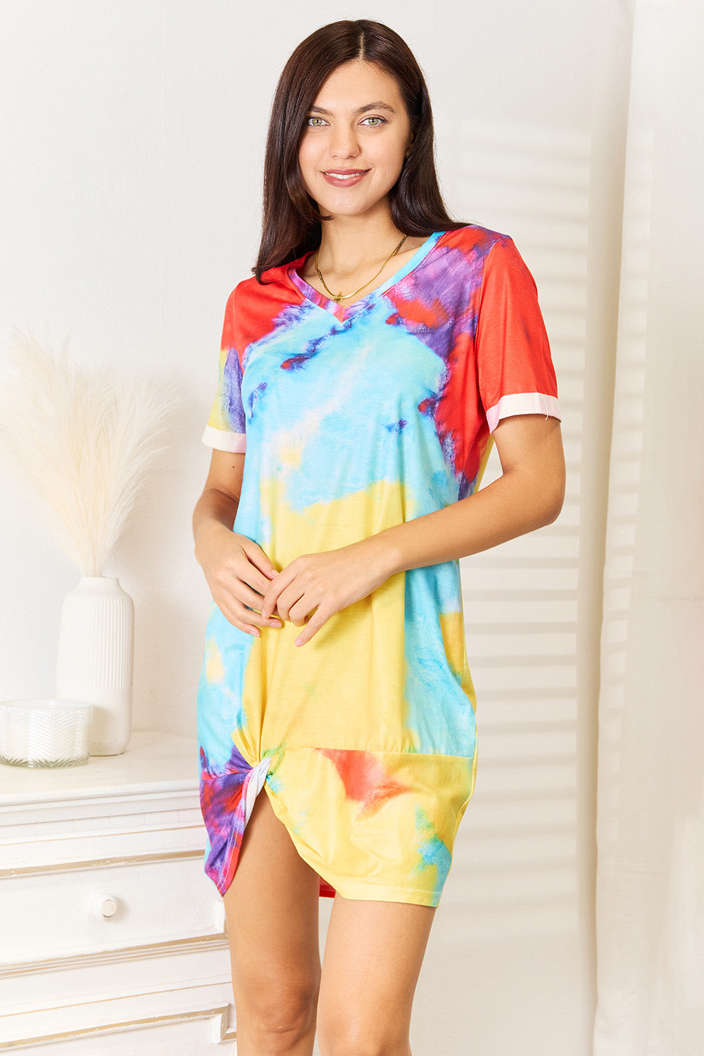 Indoors, a person with long dark hair beams as they stand in a Shiny Tie-Dye V-Neck Twisted Dress in vivid shades of blue, red, yellow, and orange. Light streams through the blinds behind them.