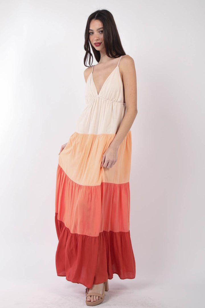 A woman in a VERY J Color Block Tiered Maxi Cami Dress with gradient cream, peach, and red tiers stands against a plain backdrop, wearing strappy sandals. Her long, straight hair flows as she looks at the camera with a slight smile.