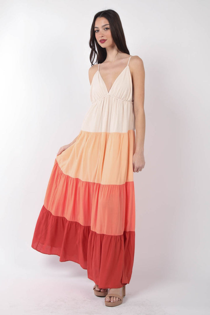 A woman against a plain white background wears the VERY J Color Block Tiered Maxi Cami Dress in shades of cream, peach, coral, and red. With long dark hair and a slight smile, she looks down while holding the flowing dress with one hand.