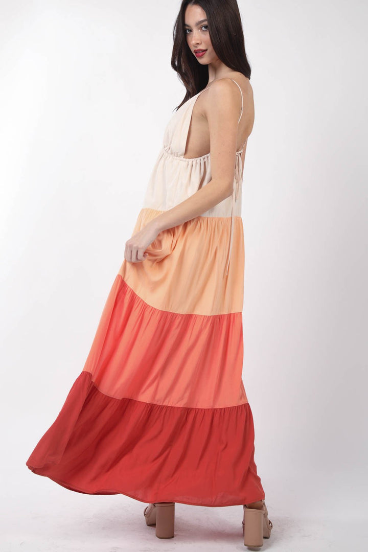 A woman with long dark hair wears the VERY J Color Block Tiered Maxi Cami Dress featuring wide horizontal stripes in cream, peach, orange, and red. Against a plain white background, she glances back over her shoulder with effortless elegance.