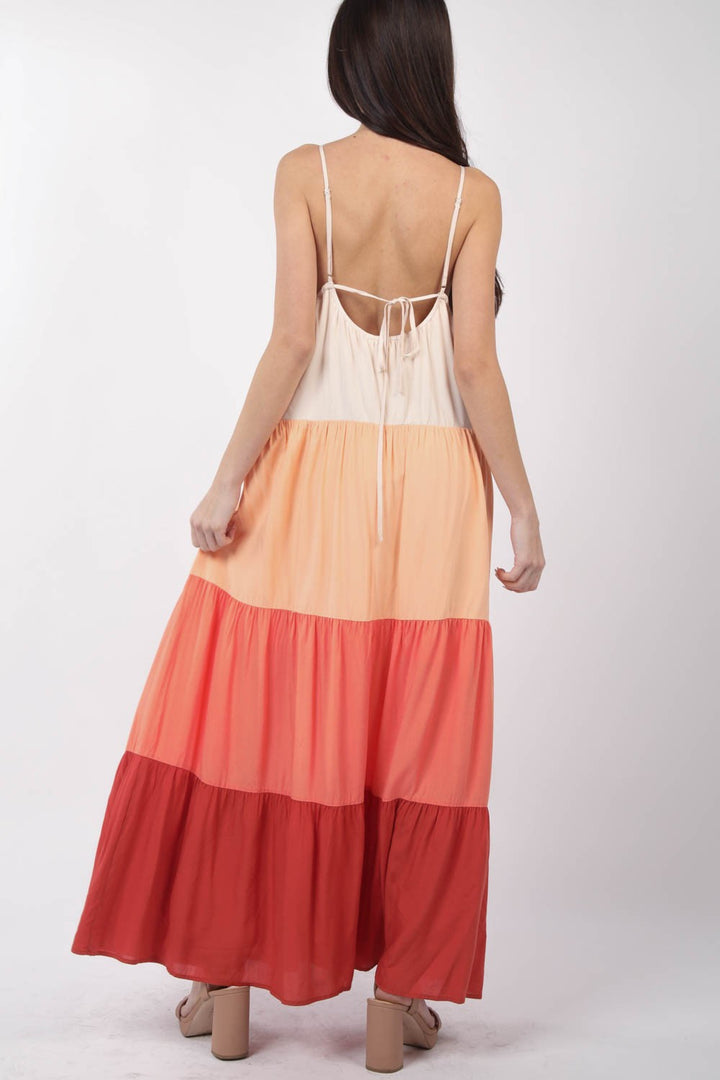 A woman with long hair wears the VERY J Color Block Tiered Maxi Cami Dress, featuring spaghetti straps and an open back, with cream, peach, coral, and red horizontal bands. She stands against a plain white background.