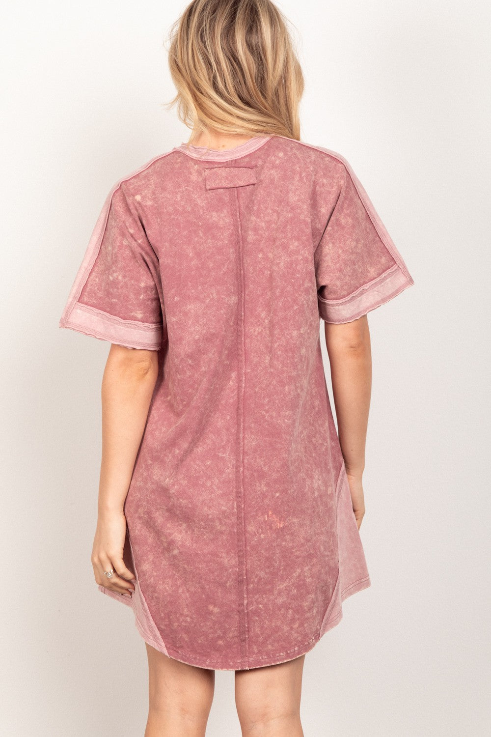 A person with long blonde hair is seen from the back wearing the VERY J Short Sleeve V-Neck Tee Dress in faded mauve, featuring visible seam details, a washed texture, and a comfortable, versatile V-neck design.