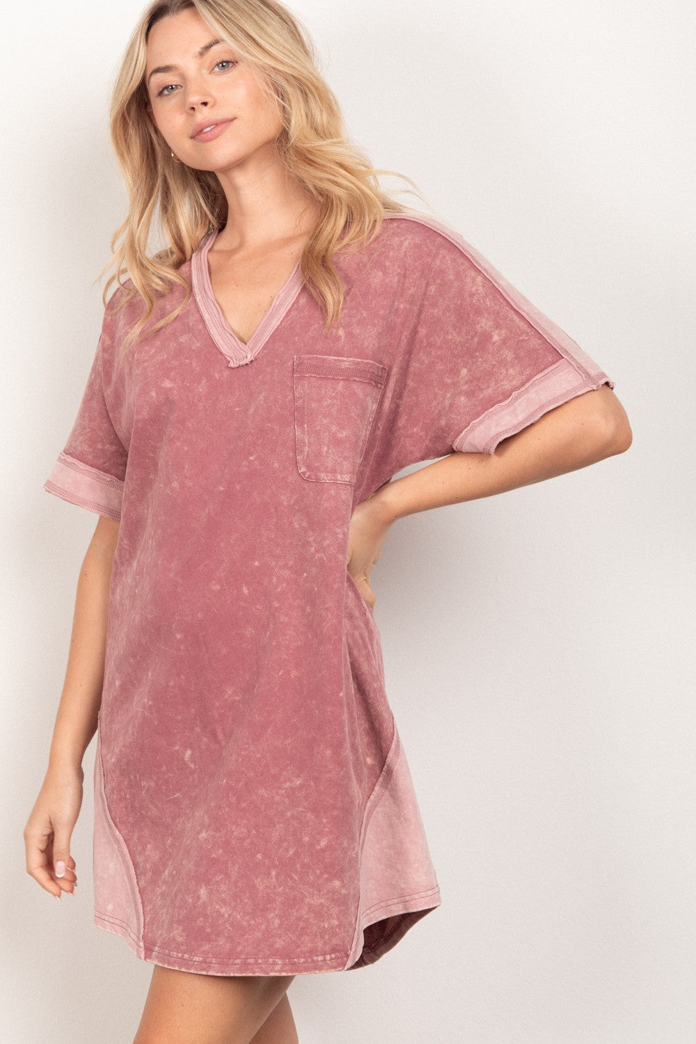 A woman with long blonde hair wears the VERY J Short Sleeve V-Neck Tee Dress in faded pink, featuring a chest pocket, as she stands against a plain light background.